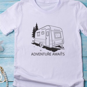 May include: White t-shirt with a black and white illustration of a caravan and the words "ADVENTURE AWAITS". The design includes a tree and grass, suggesting travel and outdoor pursuits.