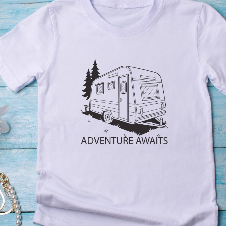 May include: White t-shirt with a black and white illustration of a caravan and the words "ADVENTURE AWAITS". The design includes a tree and grass, suggesting travel and outdoor pursuits.
