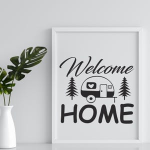 May include: A framed print with the words "Welcome Home" in black script and block letters. The design includes a camper with a heart on the side, flanked by two pine trees. A white vase with green leaves is on the left.