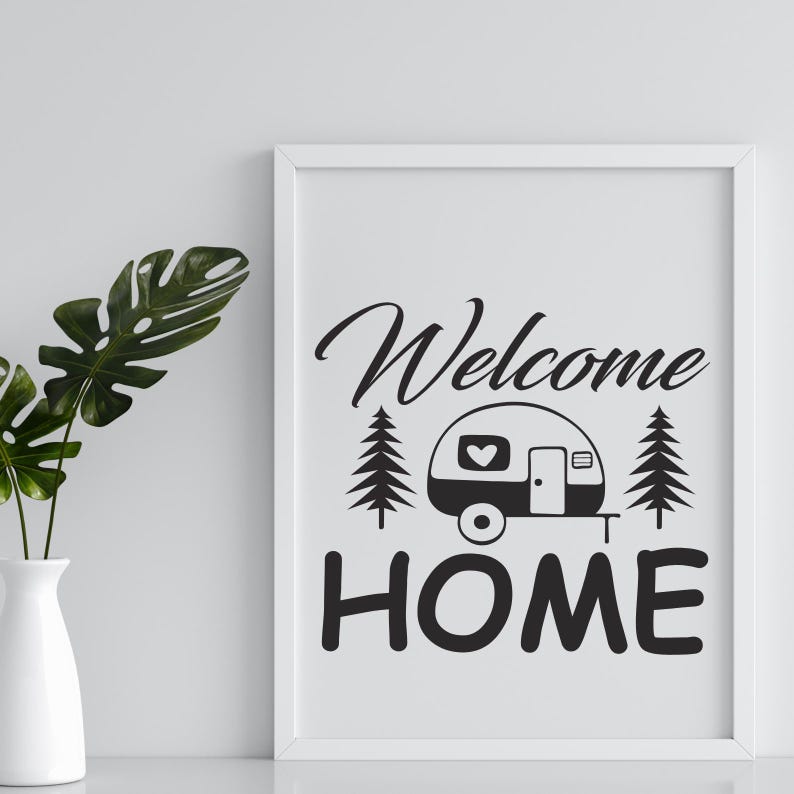 May include: A framed print with the words "Welcome Home" in black script and block letters. The design includes a camper with a heart on the side, flanked by two pine trees. A white vase with green leaves is on the left.