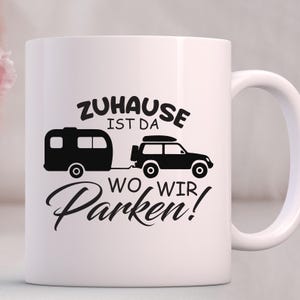 May include: White ceramic mug featuring a black graphic of a caravan and a 4x4 vehicle, connected by a tow bar. The mug has the text "Zuhause ist da wo wir Parken!"