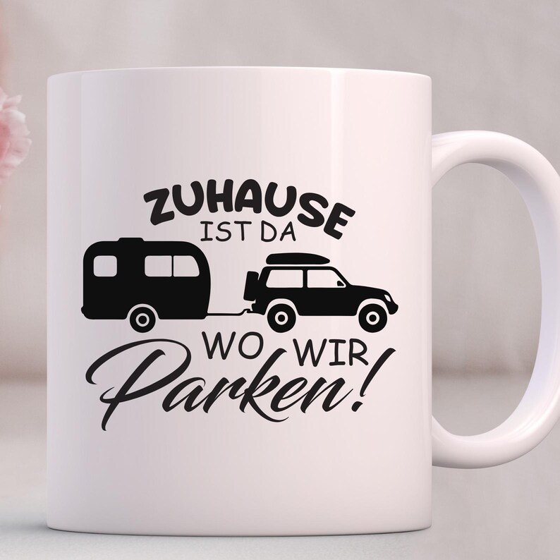 May include: White ceramic mug featuring a black graphic of a caravan and a 4x4 vehicle, connected by a tow bar. The mug has the text "Zuhause ist da wo wir Parken!"