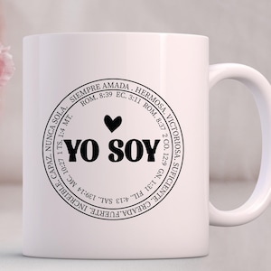 May include: White ceramic mug with a black circle design featuring the text "YO SOY" in bold letters, a heart symbol, and a circular border with inspirational text in Spanish.