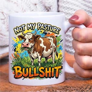 Pode incluir: Uma caneca de cer&acirc;mica branca com uma ilustra&ccedil;&atilde;o colorida de uma vaca castanha e branca em p&eacute; na relva verde. O texto acima da vaca diz NOT MY PASTURE e o texto abaixo da vaca diz BULLSHIT em letras a negrito cor de laranja e amarelo.