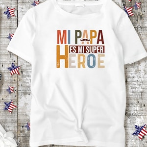 Puede incluir: Camiseta blanca con un gr&aacute;fico colorido que dice "Mi Papa Es Mi Super Heroe" con un peque&ntilde;o gr&aacute;fico de bigote sobre la palabra "Papa".