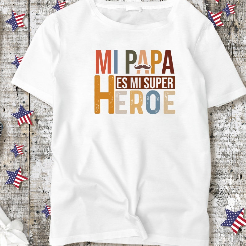 Puede incluir: Camiseta blanca con un gr&aacute;fico colorido que dice "Mi Papa Es Mi Super Heroe" con un peque&ntilde;o gr&aacute;fico de bigote sobre la palabra "Papa".