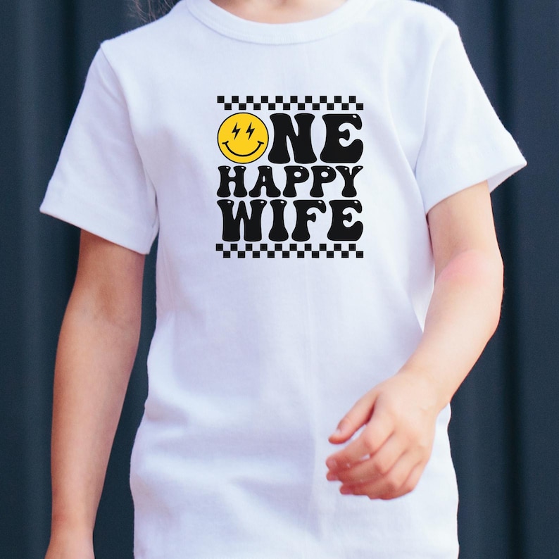 Puede incluir: Camiseta blanca con un borde de cuadros negros y blancos alrededor de una cara sonriente amarilla con rayos y el texto "ONE HAPPY WIFE" en negro.