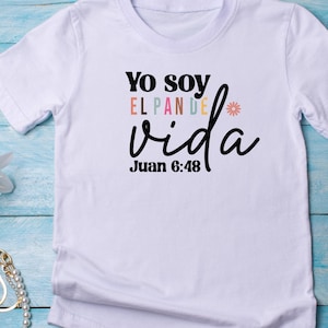 May include: White t-shirt with the text "Yo soy el pan de vida" in black with a rainbow colored "el pan de" and a small flower graphic. The text "Juan 6:48" is below the main text.
