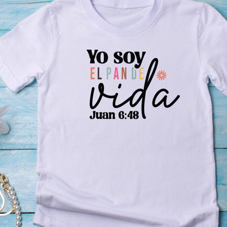 May include: White t-shirt with the text "Yo soy el pan de vida" in black with a rainbow colored "el pan de" and a small flower graphic. The text "Juan 6:48" is below the main text.