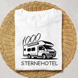 May include: White t-shirt featuring a black camper van graphic and the words "1000 STERNEHOTEL". The shirt is folded on a woven, circular, light brown surface.