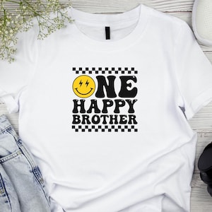 Puede incluir: Camiseta blanca con un borde de cuadros negros y blancos alrededor del texto "ONE HAPPY BROTHER" en una fuente retro. Una cara sonriente amarilla con rayos para los ojos est&aacute; por encima del texto.