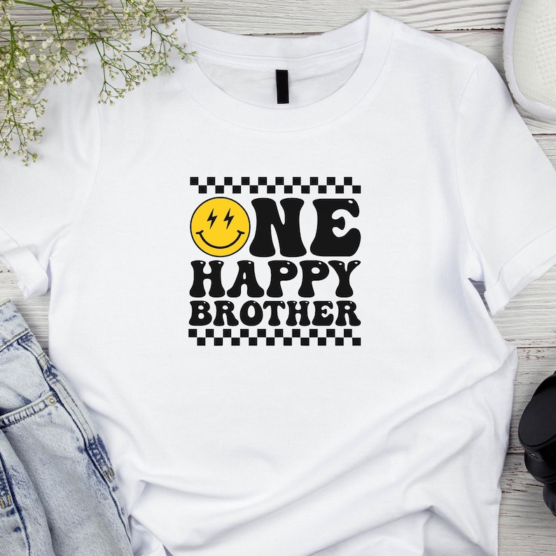 Puede incluir: Camiseta blanca con un borde de cuadros negros y blancos alrededor del texto "ONE HAPPY BROTHER" en una fuente retro. Una cara sonriente amarilla con rayos para los ojos est&aacute; por encima del texto.