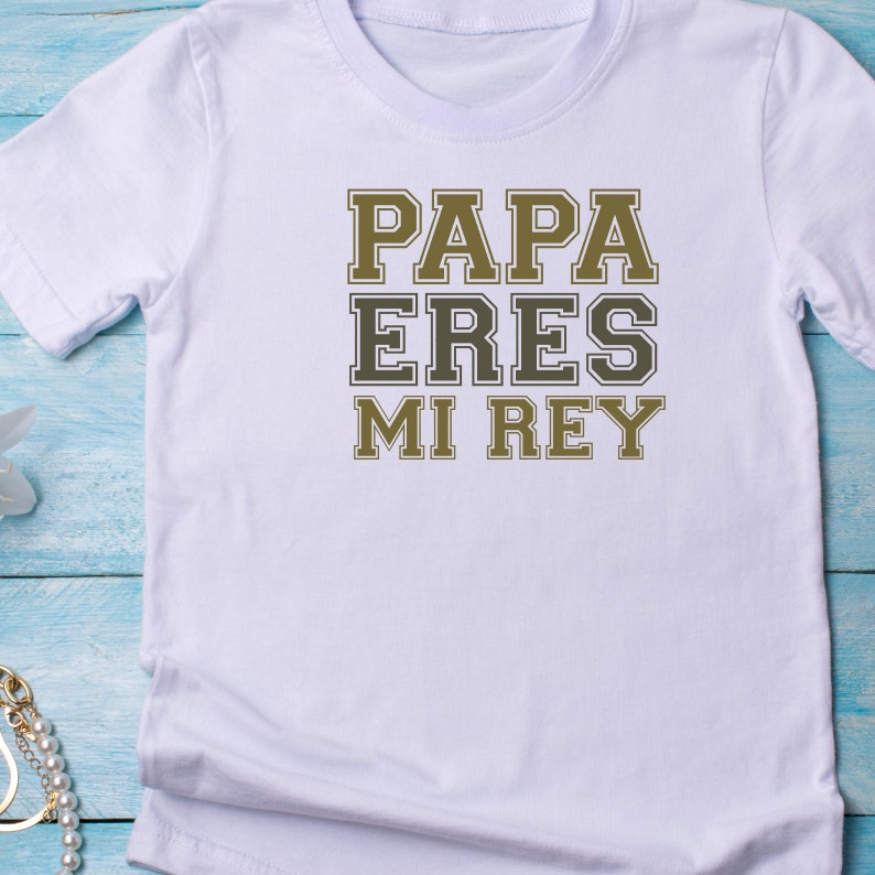 Puede incluir: Camiseta blanca con el texto "PAPA ERES MI REY" en letras negras y doradas.