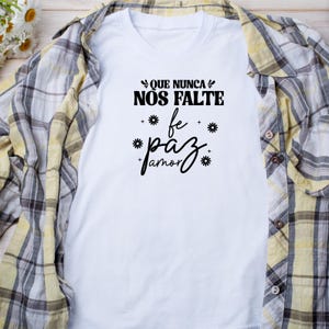 May include: White t-shirt with black text that reads "Que nunca nos falte fe paz amor" with stylized flowers around the words.