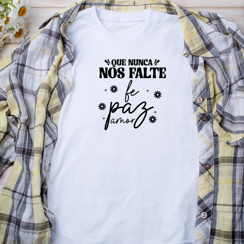 May include: White t-shirt with black text that reads "Que nunca nos falte fe paz amor" with stylized flowers around the words.