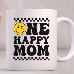 Puede incluir: Taza de caf&eacute; de cer&aacute;mica blanca con un dise&ntilde;o en negro y amarillo. La taza presenta una cara sonriente con rayos para los ojos y el texto "ONE HAPPY MOM" en una fuente retro.