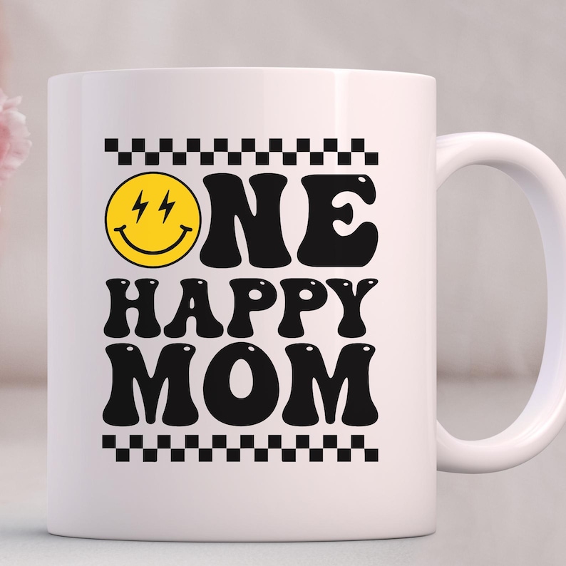 Puede incluir: Taza de caf&eacute; de cer&aacute;mica blanca con un dise&ntilde;o en negro y amarillo. La taza presenta una cara sonriente con rayos para los ojos y el texto "ONE HAPPY MOM" en una fuente retro.