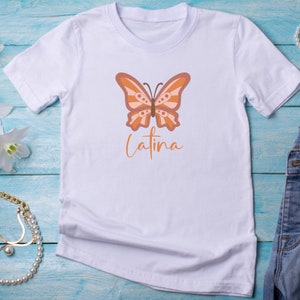 Op de afbeelding: Wit T-shirt met een bruine en oranje vlinderprint en het woord "Latina" eronder.