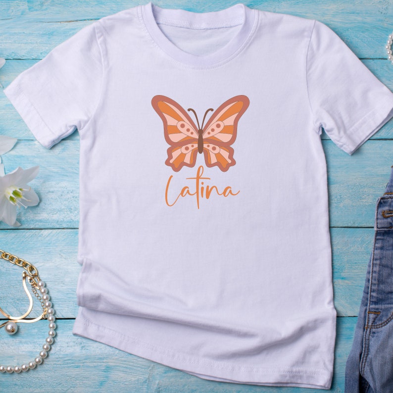 Op de afbeelding: Wit T-shirt met een bruine en oranje vlinderprint en het woord "Latina" eronder.