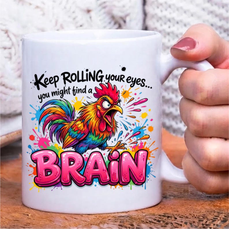 Pode incluir: Uma caneca de cer&acirc;mica branca com uma ilustra&ccedil;&atilde;o colorida de um galo com penas de arco-&iacute;ris. O texto "Keep ROLLING your eyes... you might find a BRAIN" est&aacute; em letras pretas e rosas, rodeado por salpicos de tinta vibrantes.