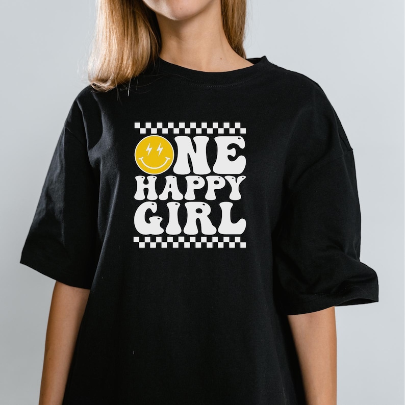 Puede incluir: Camiseta negra con un gr&aacute;fico blanco que dice "ONE HAPPY GIRL" con una cara sonriente amarilla con rayos para los ojos.