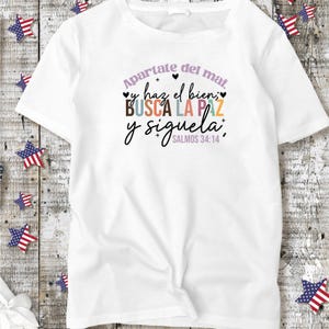 May include: White t-shirt with a colorful graphic print that says "Apartate del mal, y haz el bien; BUSCA LA PAZ y siguela." with the scripture reference "SALMOS 34:14" underneath.