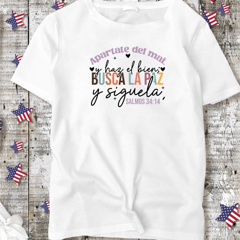 May include: White t-shirt with a colorful graphic print that says "Apartate del mal, y haz el bien; BUSCA LA PAZ y siguela." with the scripture reference "SALMOS 34:14" underneath.