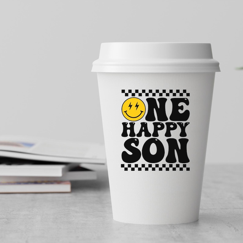Puede incluir: Vaso de caf&eacute; blanco para llevar con un borde de cuadros negros y blancos alrededor del texto "ONE HAPPY SON" con una cara sonriente amarilla con rayos para los ojos.