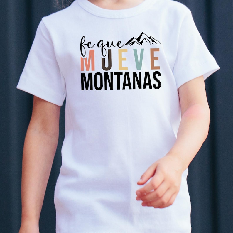 May include: White t-shirt with the text "fe que mueve montanas" in black with a rainbow color gradient for the word "mueve" and a black silhouette of mountains above the text.