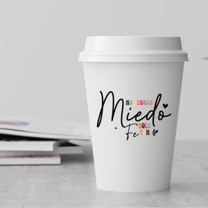 May include: White to-go coffee cup with the text "No tengas miedo + Fe solo" in colorful letters.