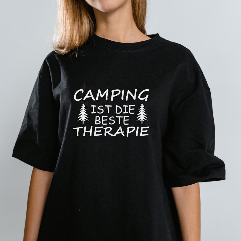 May include: Black t-shirt with the white text "CAMPING IST DIE BESTE THERAPIE" and two stylized pine trees. The shirt has short sleeves and a relaxed fit.