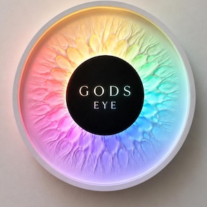 Könnte beinhalten: Rundes Kunstwerk mit weißem Rahmen, das eine farbenfrohe Regenbogen-Iris darstellt. In der Mitte befindet sich ein schwarzer Kreis mit dem Text "GODS EYE". Das Iris-Design hat eine strukturierte Oberfläche.