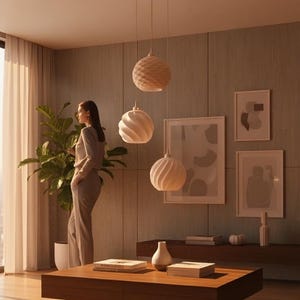 Könnte beinhalten: Innenraum mit drei Pendelleuchten mit strukturierten, kugelförmigen Schirmen. Eine Frau steht in der Nähe einer großen Pflanze und eines Fensters. Ein Holztisch und Kunstwerke sind ebenfalls zu sehen. Der Raum hat eine warme Atmosphäre.