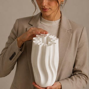 Könnte beinhalten: Eine matte, weiße Keramikvase mit einem hohen, zylindrischen Körper und vertikalen, wellenförmigen Falten, die eine gerüschte, gewellte Öffnung an der Oberseite bilden. Die Oberfläche ist glatt und einheitlich gefärbt und zeigt ein skulpturales Design.