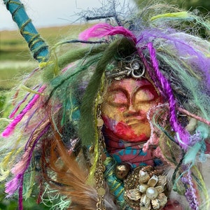 May include: A colourful, handmade doll with a face made of painted wood. The doll is adorned with feathers, beads, and yarn in shades of pink, purple, green, and gold.