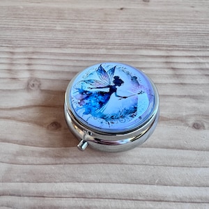 Op de afbeelding: Een ronde, zilveren pillendoos met een blauw en wit feeënontwerp op het deksel. De fee wordt afgebeeld met uitgespreide vleugels en een toverstaf tegen een sterrenachtergrond. De pillendoos heeft een kleine, zilveren knop om te openen.