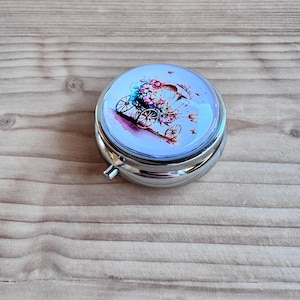 Op de afbeelding: Een ronde, zilverkleurige pillendoos met een bloemenontwerp op het deksel. Het ontwerp toont een aquarel-achtige afbeelding van een koets gevuld met bloemen in roze, paarse en blauwe tinten. De pillendoos heeft een kleine, zilveren sluiting.