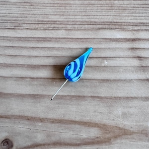 Peut inclure: Une perle en verre bleue et blanche en forme de goutte d'eau, fixée sur une épingle argentée. La perle présente un motif tourbillonnant de bleu clair et foncé. L'épingle est conçue pour la fabrication de bijoux ou comme élément décoratif.