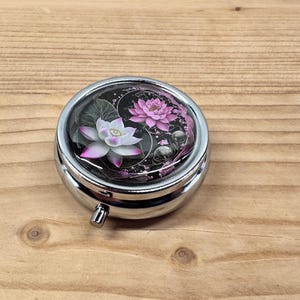 Op de afbeelding: Een ronde, zilverkleurige pillendoos met een bloemenontwerp. De bovenkant van de doos heeft een heldere, gewelfde deksel over een ontwerp van witte en roze lotusbloemen tegen een donkere achtergrond. De doos staat op een houten oppervlak.