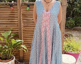 May include: A woman wearing a sleeveless blue and white patterned maxi dress with a pink and white geometric design on the side. She is standing on a white rug with a wooden floor behind her.