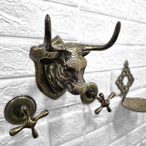 Può includere: Accessori da bagno in ottone antico montati a parete. Include un gancio a forma di testa di toro, due pomelli a forma di croce e un portasapone. La testa di toro presenta intagli dettagliati e corna curve.