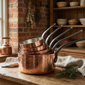 Puede incluir: Un conjunto de ollas y sartenes de cobre con asas de metal oscuro. La batería de cocina de cobre martillado se exhibe sobre una mesa de madera, con una pared de ladrillo y una ventana al fondo. Una tetera de cobre está en el alféizar.