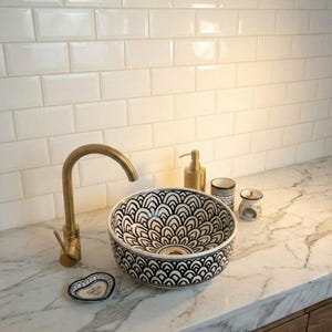 May include: A patterned bathroom sink with a black and white design. The sink is set on a marble countertop with a gold tap. Other accessories include a soap dispenser, two small cups, and a soap dish.