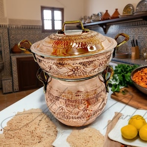 May include: A two-piece copper cooking pot with a lid, featuring intricate floral patterns. The pot has brass handles and sits on a white surface. The kitchen setting includes a wooden cutting board with food and a bowl of lemons.