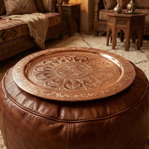 May include: A round, copper-colored metal tray with intricate embossed floral patterns rests on a brown leather ottoman. The ottoman is circular and has a stitched design. The tray is a decorative accent piece.