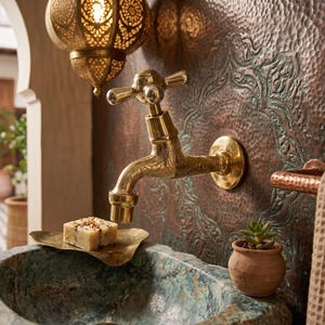 May include: A gold-toned faucet with cross handles is mounted on a textured copper wall. A small, square bar of soap rests on a brass dish above a mottled green sink. A small potted plant and a decorative hanging lamp are also visible.