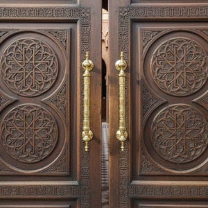May include: Intricately carved wooden double doors with gold-coloured handles. The doors are a deep brown and feature detailed geometric patterns. The design suggests a historical or cultural theme, possibly from the Middle East.