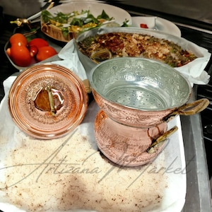 Pode incluir: Uma panela de cobre com tampa e coador, ao lado de um prato de tomates e uma bandeja de comida cozida. A panela tem detalhes ornamentados e alças de madeira. A tampa está aberta, revelando um líquido e uma guarnição.