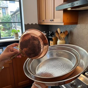 May include: A set of copper and stainless steel cookware. The copper pan has a hammered texture and a long handle. The set includes several stainless steel pans of varying sizes, perfect for cooking.
