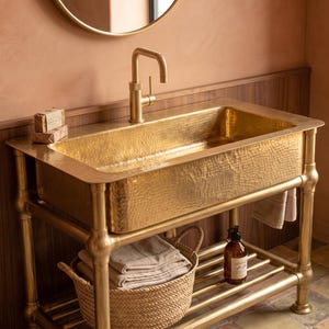 Puede incluir: Un lavabo rectangular de latón martillado integrado en una consola de metal dorado con grifo a juego. La consola cuenta con un estante inferior de listones que sostiene una cesta tejida con ropa doblada y una botella de vidrio.